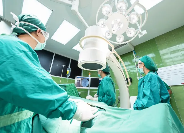 Műtőben zöld sebészi ruhát, sapkát és maszkot viselő orvosi csapat dolgozik egy páciensen műtőlámpák alatt, a háttérben monitorokon röntgenfelvétel látható.