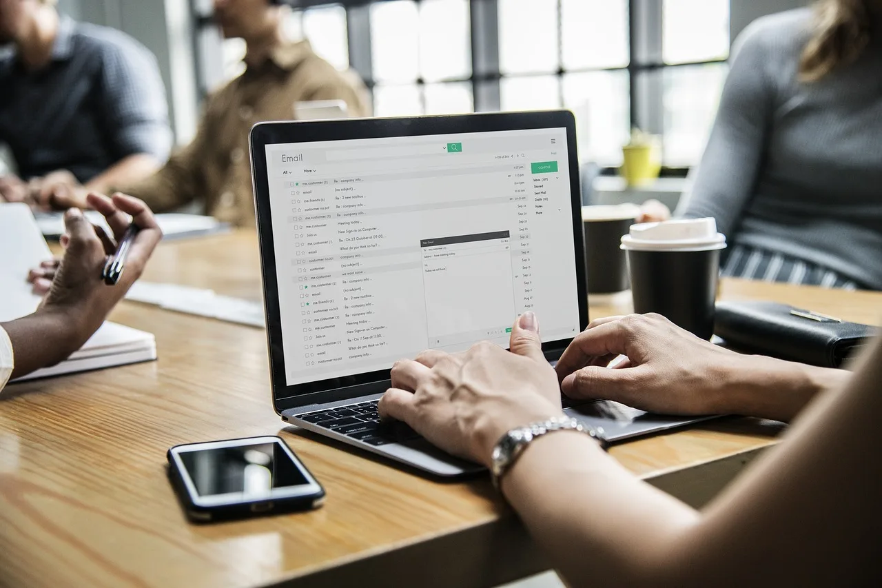 Egy asztal körül ülő embercsoport látható, előtérben egy laptop, melynek képernyőjén e-mail postafiók nyitva, egy kéz éppen gépel, mellette okostelefon és kávéspohár az asztalon.