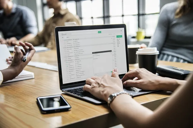 Egy asztal körül ülő embercsoport látható, előtérben egy laptop, melynek képernyőjén e-mail postafiók nyitva, egy kéz éppen gépel, mellette okostelefon és kávéspohár az asztalon.