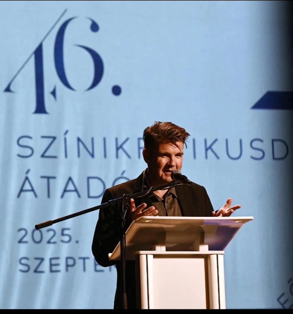 Egy férfi sötét öltönyben beszél a mikrofonba egy pulpitusnál, mögötte kék háttéren „46. Színikritikusdíj-átadó 2025. szeptember” felirat.