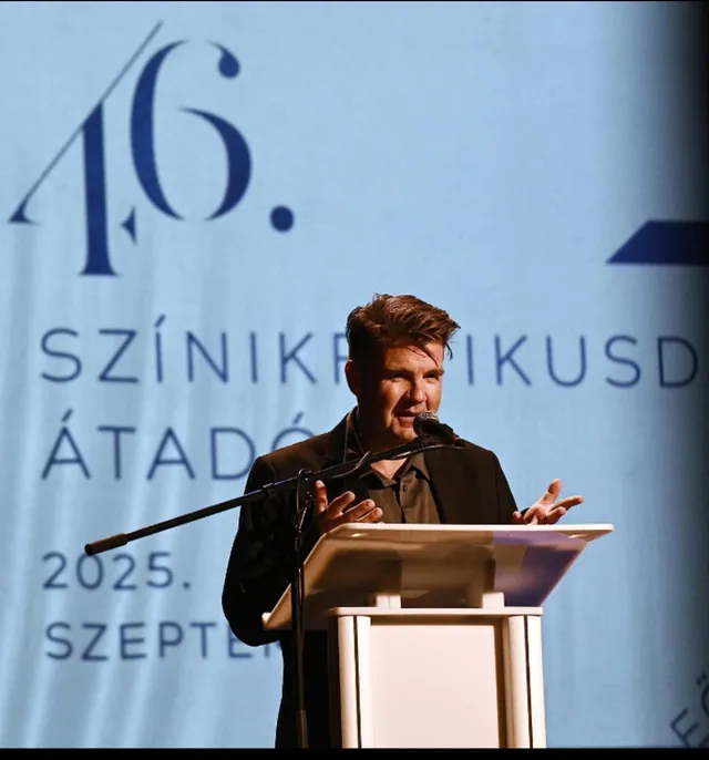 Egy férfi sötét öltönyben beszél a mikrofonba egy pulpitusnál, mögötte kék háttéren „46. Színikritikusdíj-átadó 2025. szeptember” felirat.