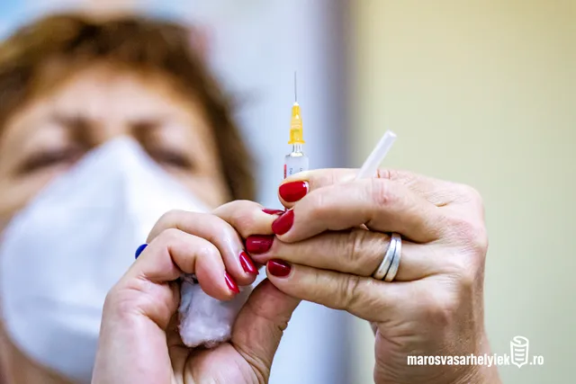 Orvosi maszkot viselő nő piros körömlakkal fest le egy ampullát, miközben egy fecskendő tűvel felszívásra előkészített injekció és egy vattacsomó látszik a kezében.