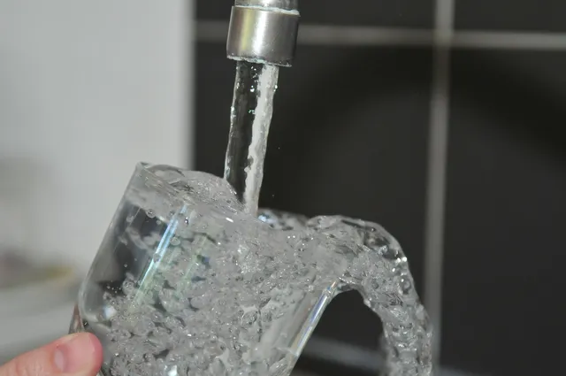 Konyhai csapból folyó víz csordul túl egy pohárból, közelről fotózva; egy kéz ujjai tartják a poharat.