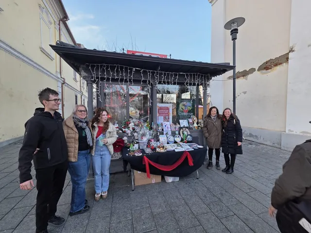 Öt fiatal felnőtt áll mosolyogva egy karácsonyi standnál, amely egy kis fabódéban van felállítva, ünnepi dekorációkkal, kézműves tárgyakkal és plakátokkal megrakott asztallal, egy régi épület előtti térkövezett utcán.
