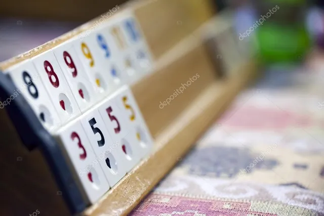 Közelkép egy fa kártyatartóról, rajta remi (rummikub) színes számkockákkal, elmosódott háttérrel.
