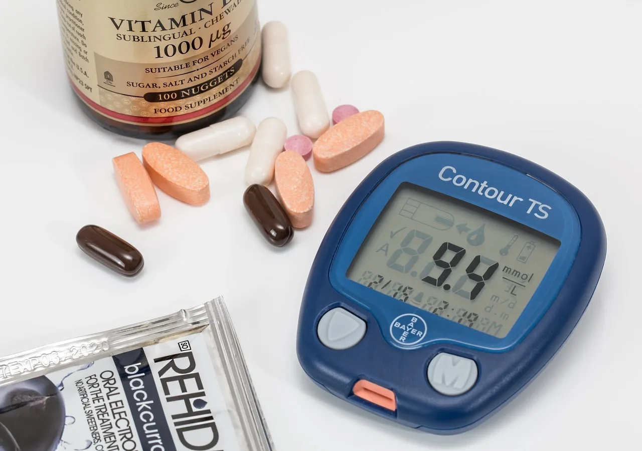 Egy vércukormérő készülék (Contour TS) 9,4 mmol/l értéket mutat a kijelzőjén, mellette különböző tabletták, kapszulák, egy vitamin étrend-kiegészítő flakon és egy elektrolit-italpor tasakja látható fehér felületen.
