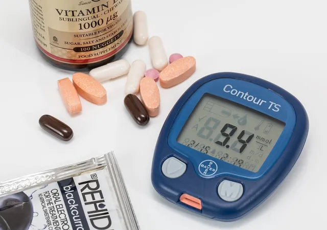 Egy vércukormérő készülék (Contour TS) 9,4 mmol/l értéket mutat a kijelzőjén, mellette különböző tabletták, kapszulák, egy vitamin étrend-kiegészítő flakon és egy elektrolit-italpor tasakja látható fehér felületen.