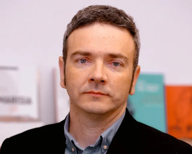 Rövid, ősz-barna hajú férfi közeli portréja, sötét zakóban és szürkéskék ingben, komoly arckifejezéssel néz a kamerába. A háttérben elmosódott könyvborítók látszanak.