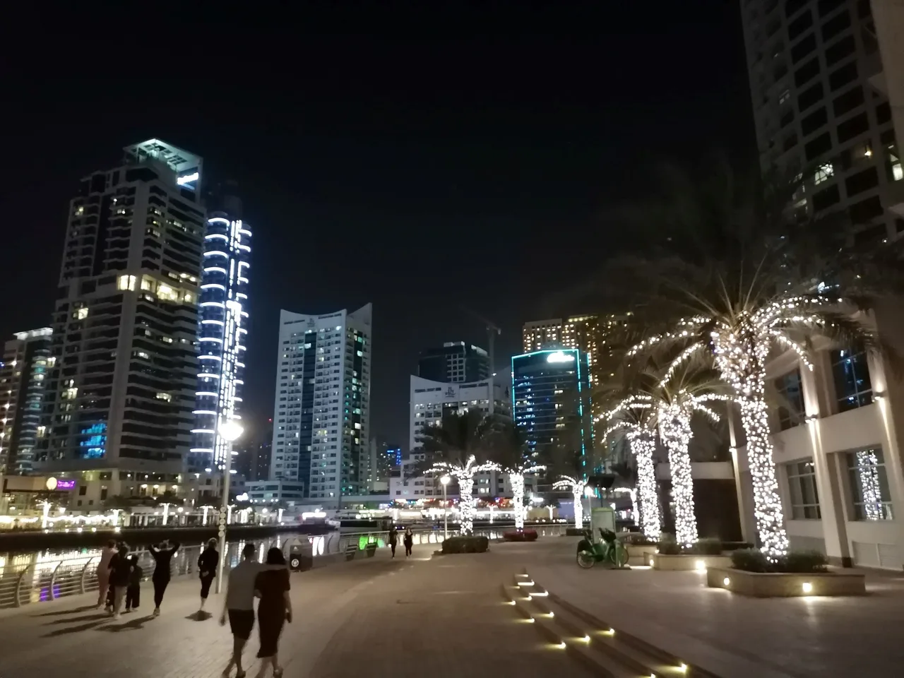Éjszakai városkép Dubajban: fényárban úszó felhőkarcolók egy csatorna mentén, jobb oldalon fénnyel díszített pálmafák, a sétányon néhány sétáló ember.