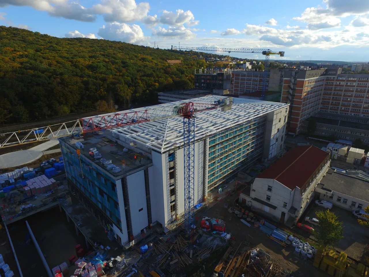 Légi felvétel egy nagy, még épülő többszintes kórházépületről, melynek homlokzatán üveg- és panelburkolat látható, tetején fém tetőszerkezet és két épületszerelő toronydaru dolgozik. A háttérben erdős domboldal, valamint meglévő kórházi tömbök és lakóépületek láthatók.