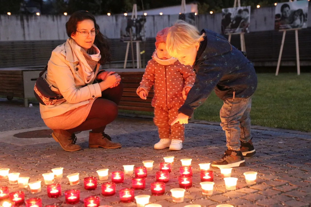 Egy nő két kisgyermekkel guggol és hajol a földre kirakott, gyertyákkal megvilágított poharak fölé esti szabadtéri rendezvényen, mögöttük fényfüzérrel díszített kerítés és kiállított fotók láthatók.