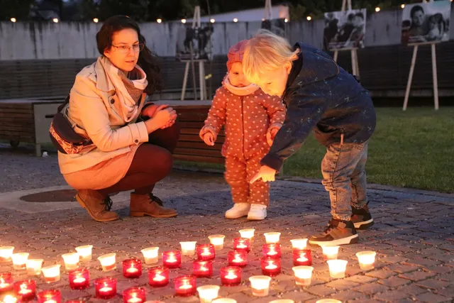 Egy nő két kisgyermekkel guggol és hajol a földre kirakott, gyertyákkal megvilágított poharak fölé esti szabadtéri rendezvényen, mögöttük fényfüzérrel díszített kerítés és kiállított fotók láthatók.