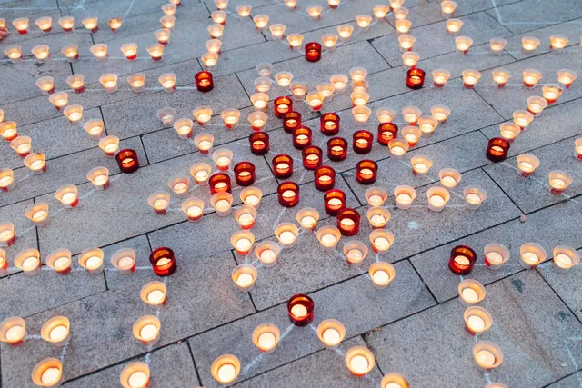 Kőburkolatú téren mintázatba rendezett gyertyák égnek, fehér és piros színű poharakban, alakzatokat és vonalakat kirajzolva a járólapokon.