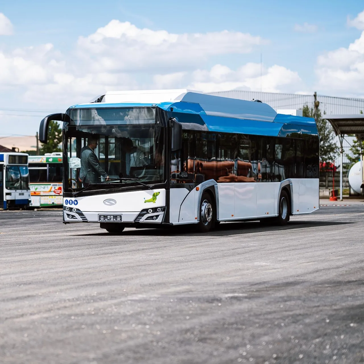Modern kék-fehér színezésű, alacsonypadlós autóbusz áll egy aszfaltozott téren, sofőrje beszáll a vezetőülésbe; háttérben régebbi buszok és ipari épületek láthatók.