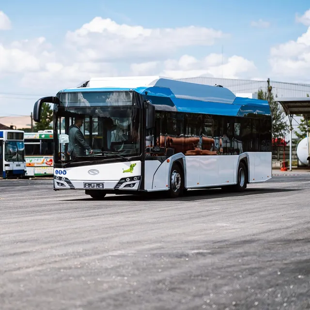 Modern kék-fehér színezésű, alacsonypadlós autóbusz áll egy aszfaltozott téren, sofőrje beszáll a vezetőülésbe; háttérben régebbi buszok és ipari épületek láthatók.
