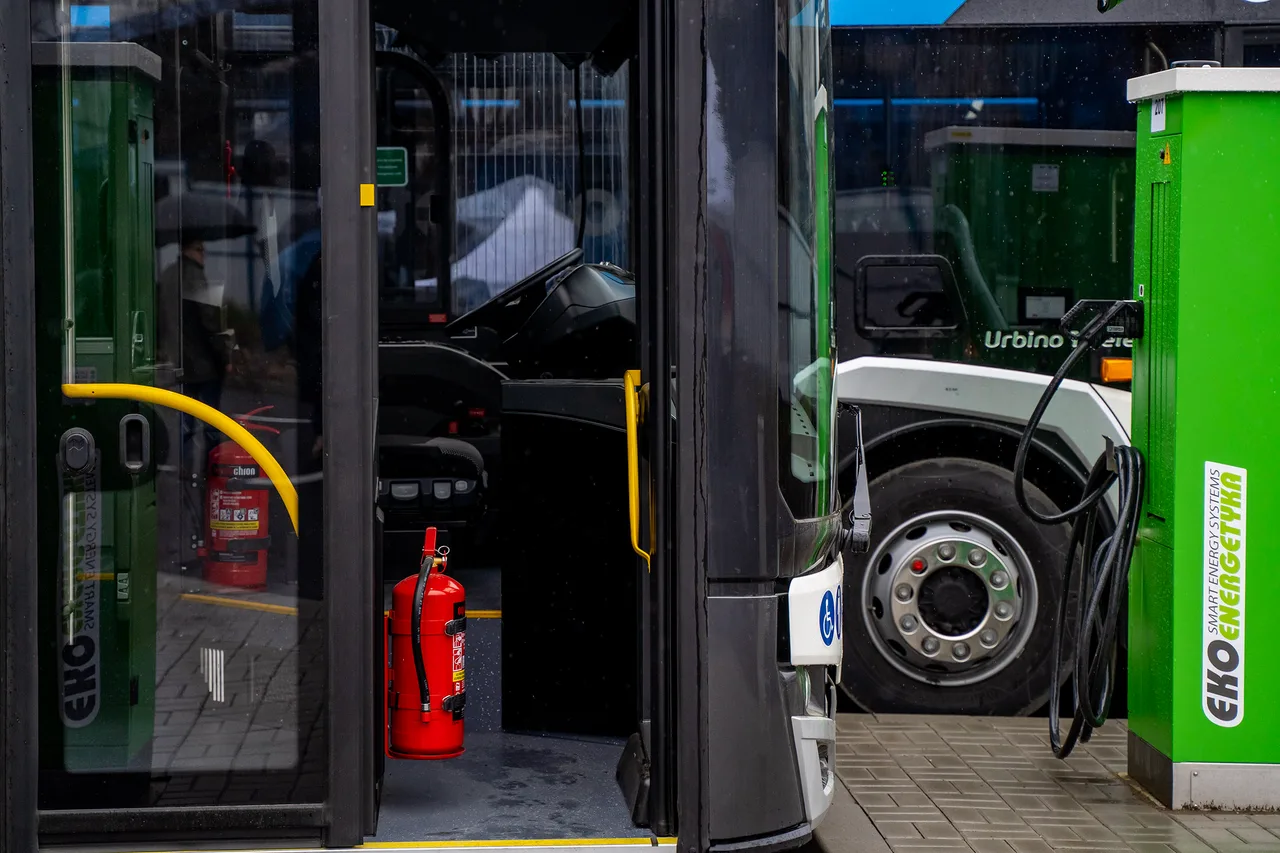 Elektromos autóbusz nyitott ajtaja látszik, belül tűzoltó készülékkel, mellette egy zöld színű elektromos töltőállomás (Eko Energetyka) van csatlakoztatva a busz elejéhez, jobbra egy másik Urbino feliratú busz áll.