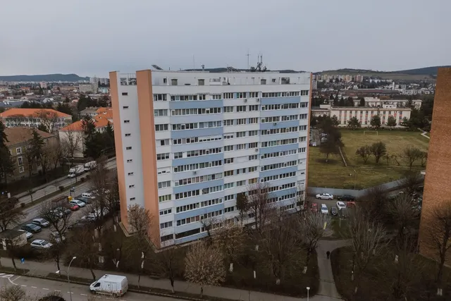 Légi felvétel egy magas, panel tömbházról, amelynek homlokzatán fehér és kék hőszigetelő panelek láthatók, két oldalán narancssárga oldalfalakkal. Körülötte parkolók, fák, és háttérben a város beépítése, valamint egy sportpálya látható.