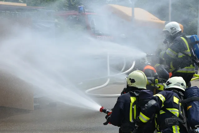 Tűzoltók sisakban és védőfelszerelésben tömlőkkel oltják a tüzet, erős vízsugarat és gőzt/füstöt bocsátva ki egy jármű irányába, mögöttük tűzoltóautó látható.