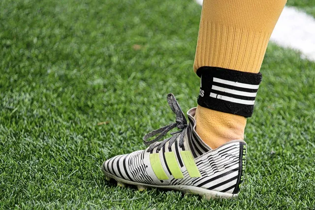 Közelkép egy focista lábáról: fehér-fekete mintás stoplis cipő zöld csíkokkal, sárga sportzokni és fekete-fehér csíkos bokaszalag a füvön.