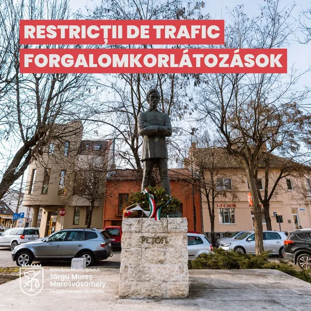 Petőfi Sándor szobra egy téren, talapzatán nemzeti színű szalaggal díszített koszorúval, körülötte parkolóautók és háttérben lakóházak. A képen felül piros sávban „RESTRICȚII DE TRAFIC / FORGALOMKORLÁTOZÁSOK” felirat olvasható.
