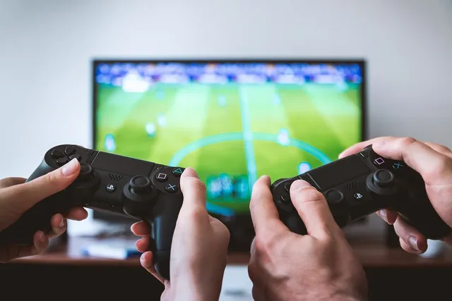 Két személy kezében PlayStation kontroller látható közelről, háttérben egy tévéképernyőn futballvideojáték jelenete jelenik meg.