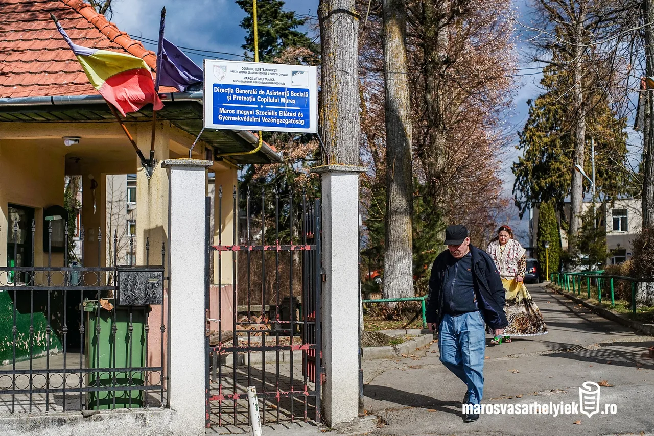 Bejárat egy épülethez, amelynek kapuján kék tábla jelzi: Maros Megyei Szociális Ellátási és Gyermekvédelmi Vezérigazgatóság. Az épület előtt román zászló lóg, a járdán egy idős férfi és egy idős nő sétál el a kapu mellett.