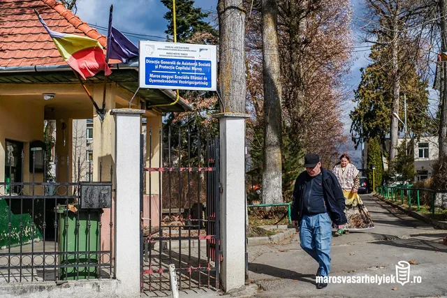 Bejárat egy épülethez, amelynek kapuján kék tábla jelzi: Maros Megyei Szociális Ellátási és Gyermekvédelmi Vezérigazgatóság. Az épület előtt román zászló lóg, a járdán egy idős férfi és egy idős nő sétál el a kapu mellett.