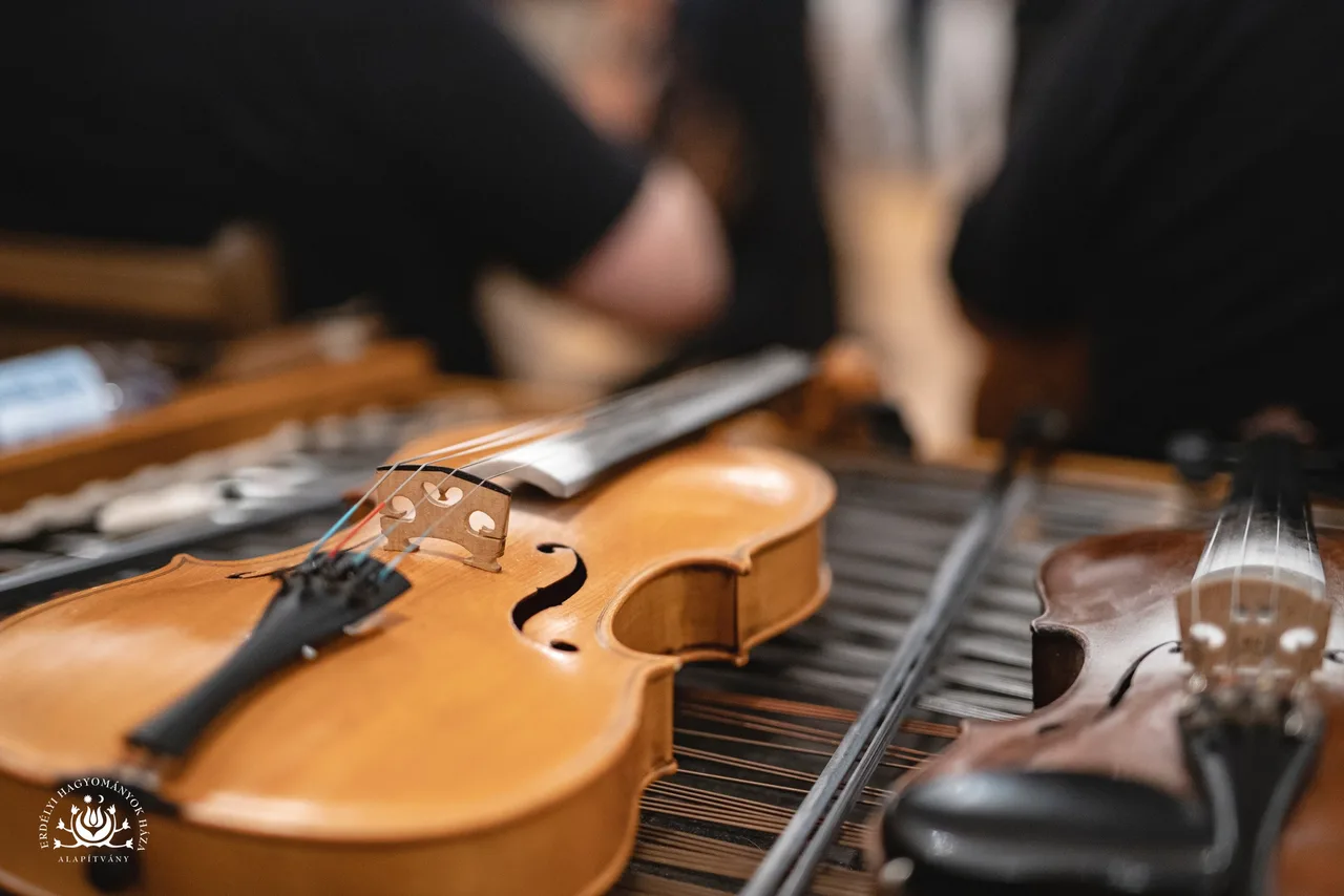 Közelről fotózott hegedűk cimbalom felett, előtérben egy hegedű teste és vonója, jobb oldalon egy másik hegedű nyaka látszik, háttérben elmosódottan emberek kezei.