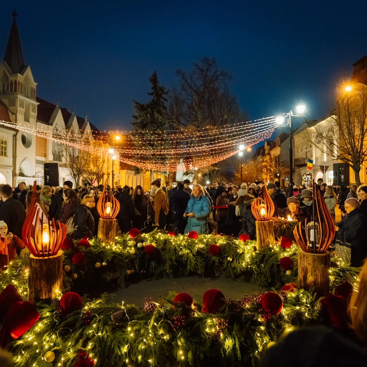 Esti utcai jelenet egy téren, ahol óriási, fenyőágakból és piros díszekből font adventi koszorú áll, gyertya alakú fa díszekkel és fényfüzérekkel, körülötte sokan gyülekeznek egy templom és fényfüzérekkel átszőtt tér előtt.