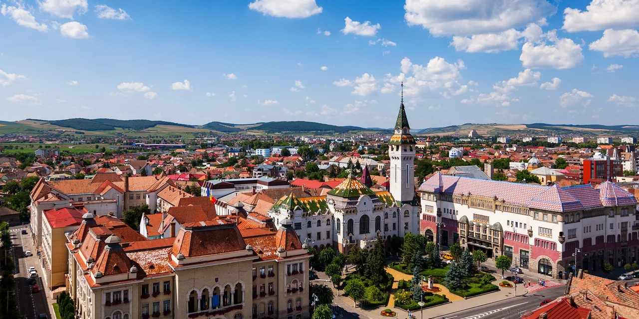 Madártávlati kép Marosvásárhely belvárosáról, középen a Kultúrpalota tornyával és a Prefektúra épületével, körülöttük cserepes háztetők, zöld park és háttérben dombok láthatók.