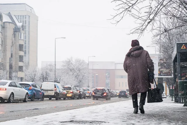 Havas utcán hátulról fotózott nő sétál vastag télikabátban, sapkában, kezében táskával, mellette forgalmas, hóban araszoló autósor és havas fák láthatók.