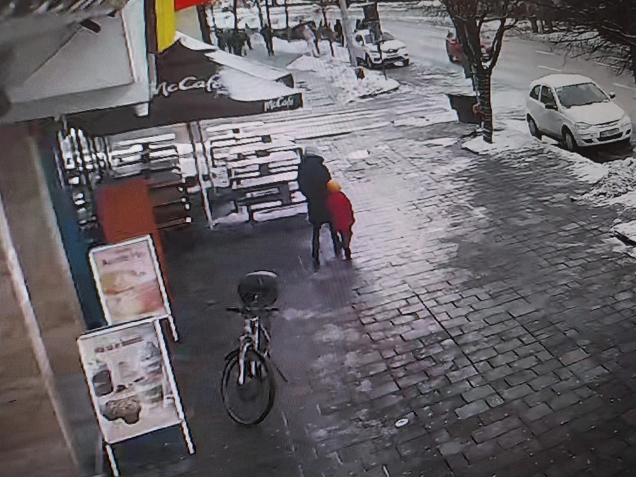 Biztonsági kamera felvétele havas, jeges utcáról egy McCafé terasza előtt, ahol két ember áll egymás mellett; a járdán kerékpár van kitámasztva, az úton hó és parkoló autók láthatók.
