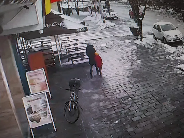 Biztonsági kamera felvétele havas, jeges utcáról egy McCafé terasza előtt, ahol két ember áll egymás mellett; a járdán kerékpár van kitámasztva, az úton hó és parkoló autók láthatók.