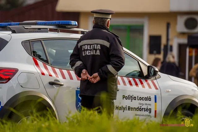 Marosvásárhelyi helyi rendőr sapkás egyenruhában, hátat fordítva, kezét háta mögött összefogva áll egy Politia Locala feliratú rendőrautó mellett.