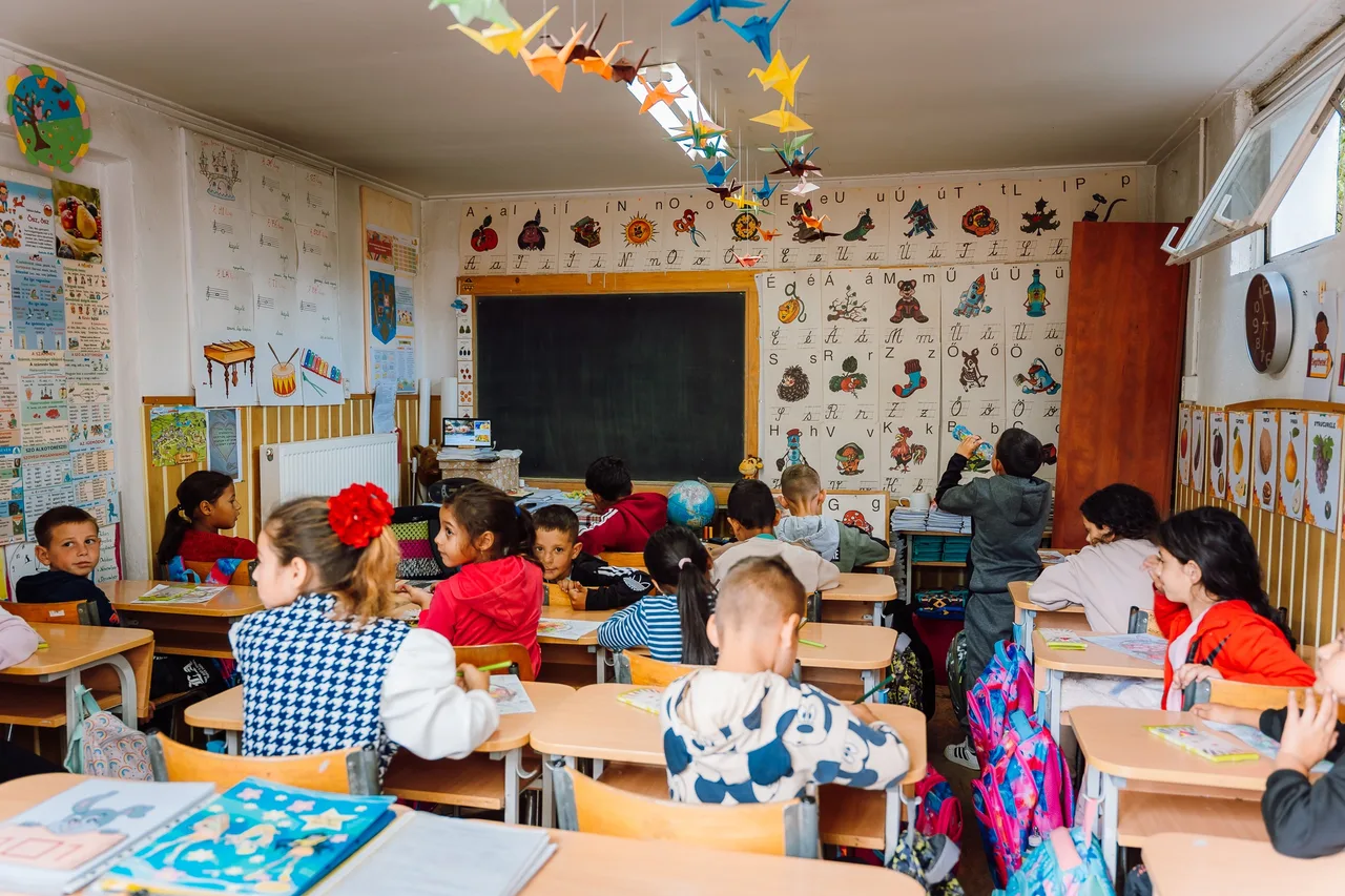 Osztályterem, ahol kisiskolás gyerekek ülnek padjaikban, a táblák felett magyar ábécés tablók és színes origami madarak lógnak a mennyezetről. Néhány gyerek dolgozatfüzetben ír, egy fiú a szekrény mellett áll.