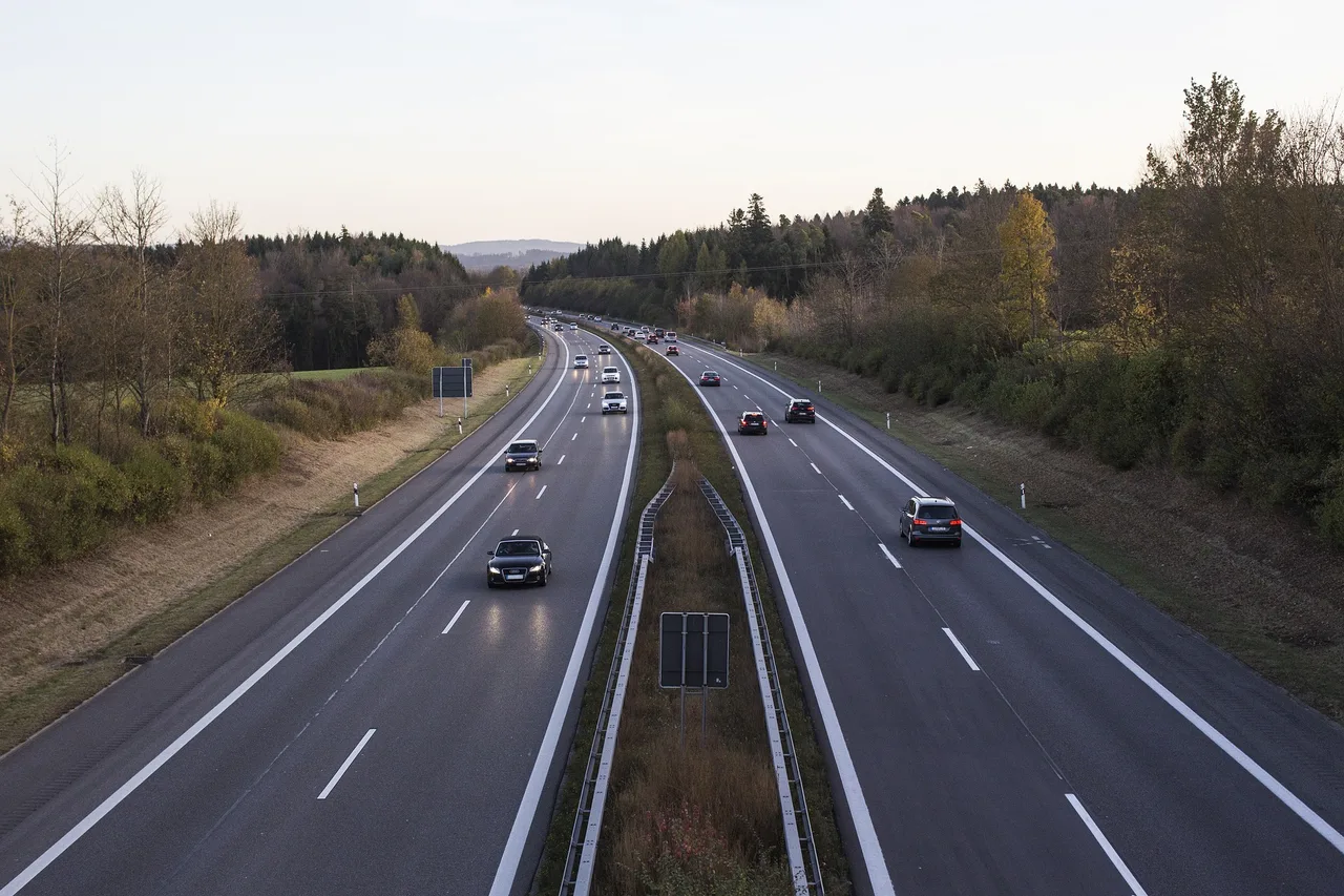 Magaslati nézet egy kétszer két sávos autópályáról, mindkét irányban több autóval, körülöttük ősziesen színes erdős domboldal.