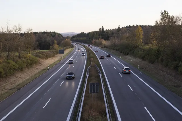 Magaslati nézet egy kétszer két sávos autópályáról, mindkét irányban több autóval, körülöttük ősziesen színes erdős domboldal.