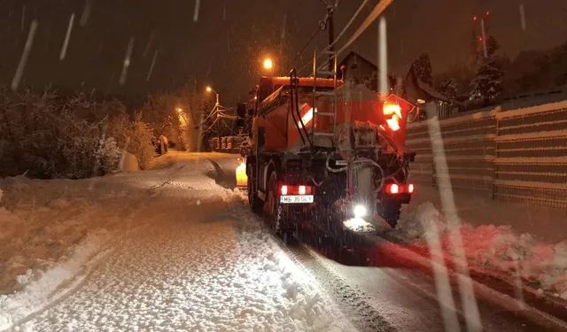 Éjszaka, havazásban egy narancssárga hóeltakarító/sószóró teherautó áll egy havas úton, lámpái világítanak, mellette kerítés és havas fák.