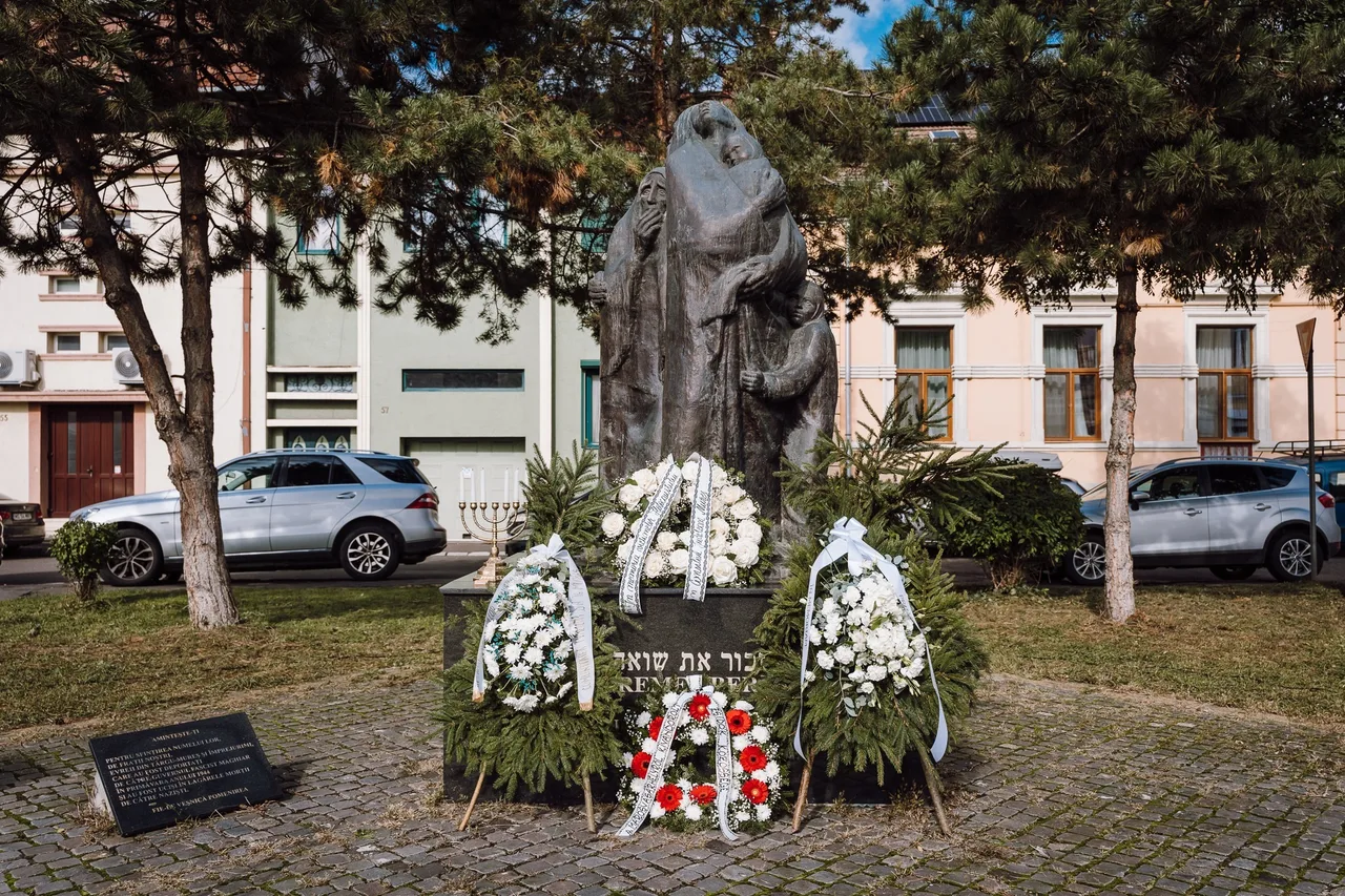 Holokauszt-emlékmű szoborcsoportja Marosvásárhelyen, alatta héber és román nyelvű felirat, körülötte fehér és piros virágból font koszorúk, mellette hétágú gyertyatartó (menóra) áll. A háttérben színes homlokzatú házak és parkoló autók láthatók.