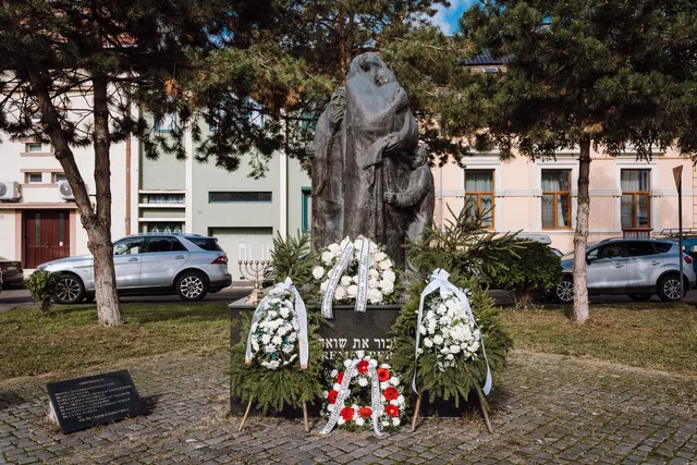 Holokauszt-emlékmű szoborcsoportja Marosvásárhelyen, alatta héber és román nyelvű felirat, körülötte fehér és piros virágból font koszorúk, mellette hétágú gyertyatartó (menóra) áll. A háttérben színes homlokzatú házak és parkoló autók láthatók.