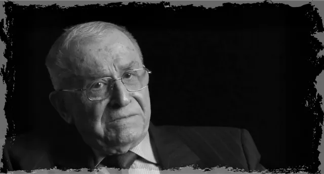 Fekete-fehér portré egy idős férfiról, aki szemüveget és sötét öltönyt visel, egyenesen a kamerába néz, komoly arckifejezéssel.