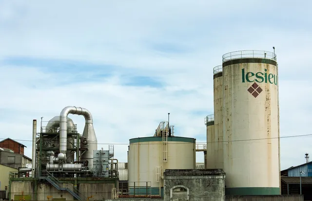 Ipari üzem tárolótartályai és csővezetékei látszanak, az egyik nagy silón „lesieu” felirat és rombusz alakú logó olvasható, felhős égbolt előtt.