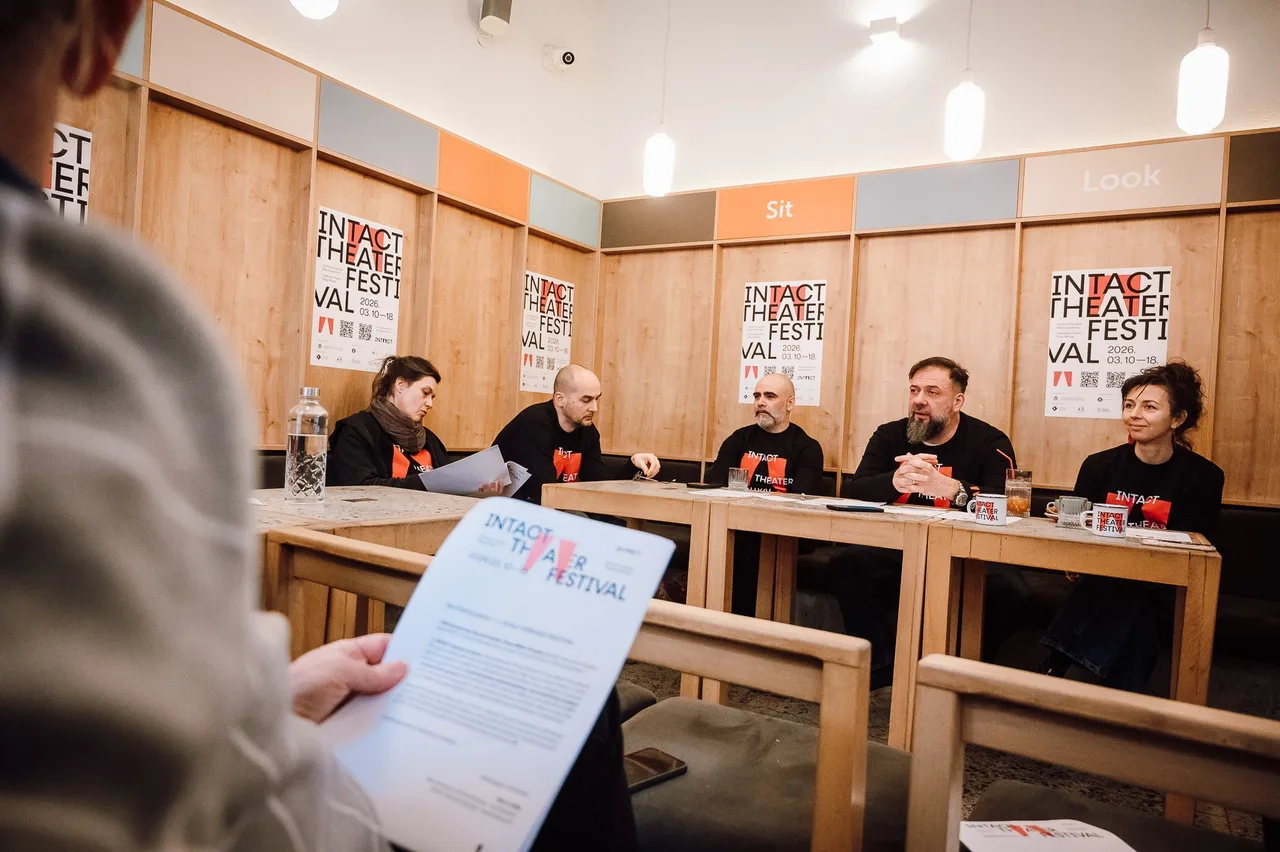 Sajtótájékoztató egy teremben, ahol öt egyforma fekete pólóban ülő ember beszélget egy asztal mögött, mögöttük az INTACT Theater Festival plakátjai láthatók a falon. Előtérben egy néző kezében a fesztivál programfüzetét tartja.