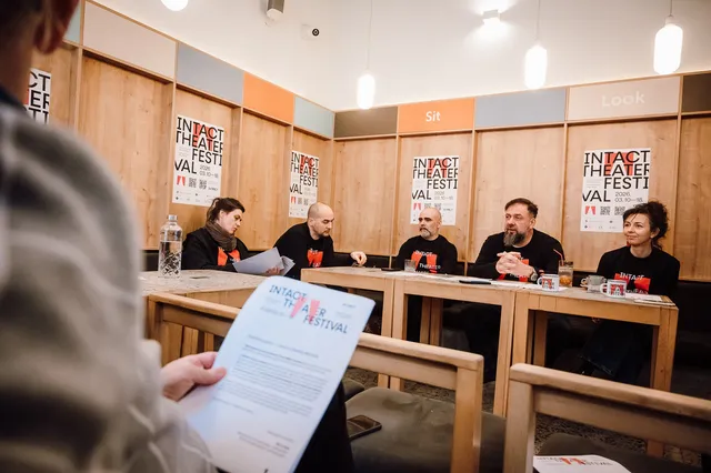 Sajtótájékoztató egy teremben, ahol öt egyforma fekete pólóban ülő ember beszélget egy asztal mögött, mögöttük az INTACT Theater Festival plakátjai láthatók a falon. Előtérben egy néző kezében a fesztivál programfüzetét tartja.