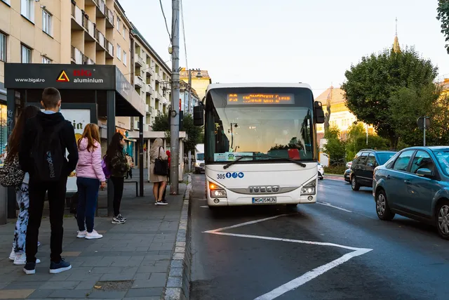 Buszmegállóban várakozó diákok láthatók hátulról, miközben egy fehér IVECO autóbusz áll meg mellettük az utcán, körülötte forgalom és parkoló autók.