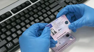 Kék gumikesztyűt viselő kezek billentyűzet előtt tartanak és vizsgálnak egy jogosítványt vagy személyazonossági igazolványt.