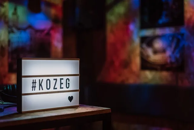 Világító feliratos doboz egy asztalon, rajta '#KOZEG' felirat és egy szív szimbólum, a háttérben elmosódott, színes fényekkel megvilágított terem.