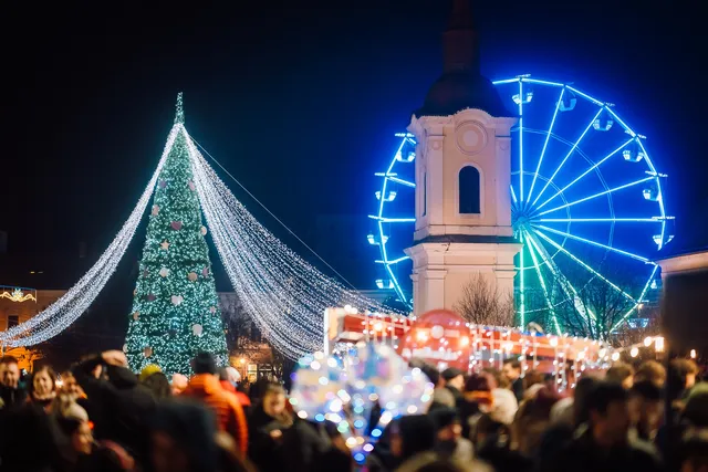 Éjszakai karácsonyi vásár térre díszített karácsonyfa, fényfüzérek és egy kék-zöld fénnyel világító óriáskerék látható, a háttérben egy templom óratornya emelkedik ki, előtérben nagy tömeg sétál a fények alatt.