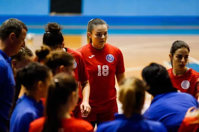 Piros mezes női kézilabdázók csapata áll körben egy sportcsarnokban, középen a 18-as számú játékos, a háttérben kék mezes ellenfelek is láthatók.