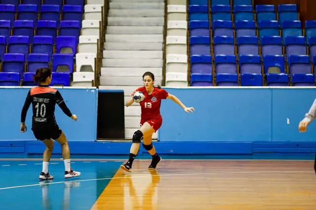 Üres lelátójú sportcsarnokban női kézilabdázók mérkőzést játszanak: piros mezes játékos labdával fut, mellette fekete-narancs mezes ellenfél védekezik.